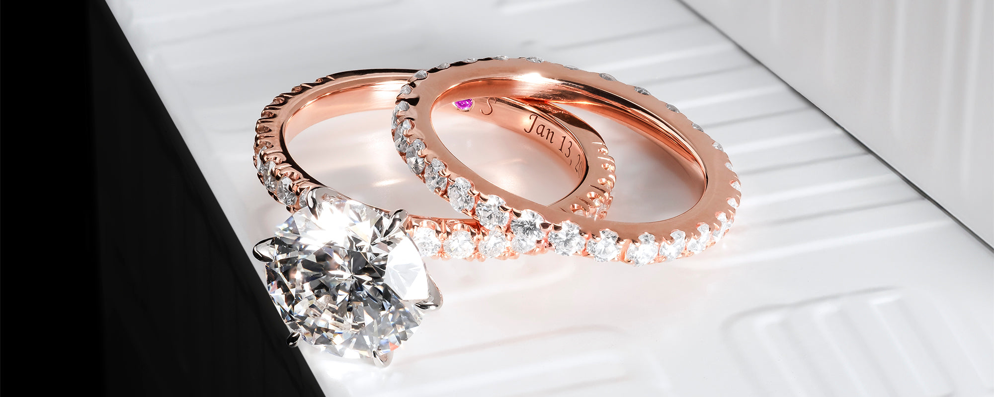 Close-up of a rose gold diamond engagement ring set featuring a round-cut solitaire engagement ring and a matching pavé diamond wedding band. Both rings are adorned with sparkling diamonds along the band, with a hidden pink diamond inside the band. The engagement ring is engraved with a date, adding a personal touch. The composition showcases the brilliance of the diamonds and the warm tones of the rose gold, ideal for those seeking a luxurious and personalised ring set.