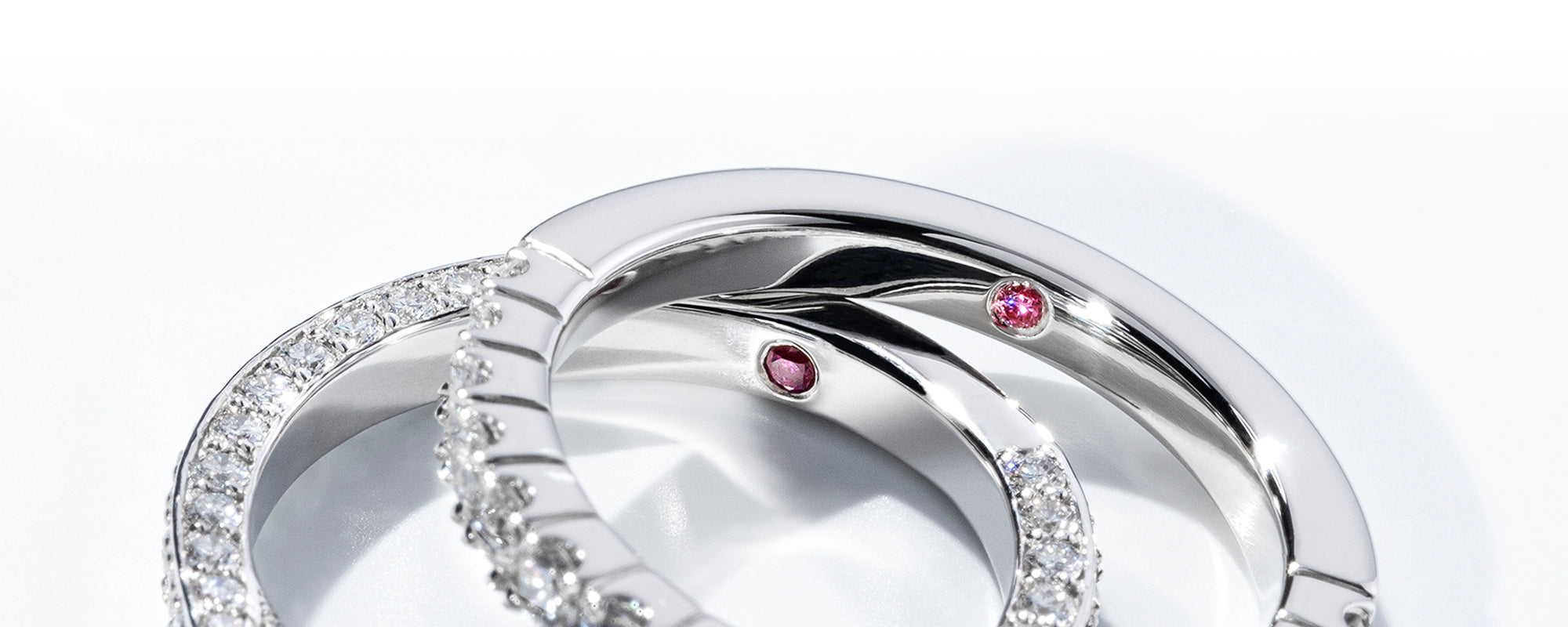 Close-up of two platinum eternity bands, each featuring a rare hidden pink diamond inside the band. The pink diamonds take centre stage, adding a unique and personal touch to both rings. One band is adorned with pavé-set diamonds, while the other has a sleek polished finish. The pink diamonds symbolise exclusivity and rarity, making these rings perfect for those seeking a luxurious, personalised design.