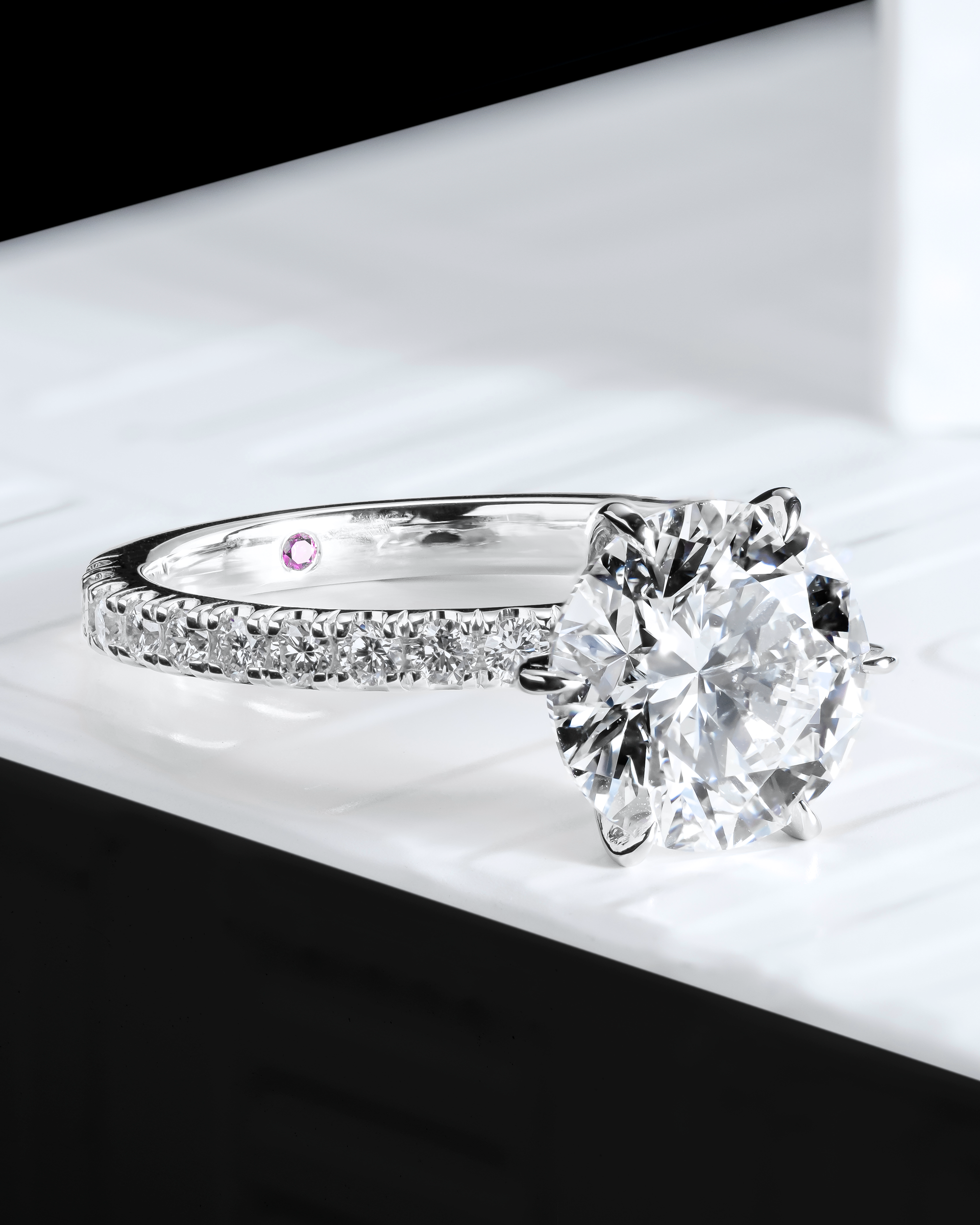 Close-up of a round brilliant-cut diamond engagement ring featuring a pavé diamond band in polished white gold. A hidden pink diamond is set inside the band, symbolising a unique and personal touch. The image highlights the brilliant sparkle of the centre stone and the intricate detailing of the band, set against a sleek, modern background. Ideal for those seeking a timeless yet distinctive engagement ring design that combines elegance and contemporary style.