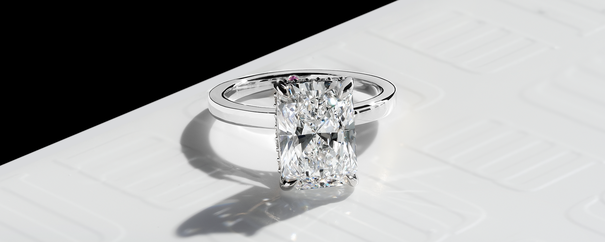 Close-up view of a radiant-cut diamond engagement ring displayed on a polished white surface with the MJ Jones monogram subtly embossed. The ring showcases a stunning radiant-cut diamond with sharp corners, reflecting brilliance and clarity. A hidden pink diamond accent is visible beneath the center stone, adding a touch of uniqueness. The composition highlights the diamond’s clean facets and sparkle, complemented by a sleek & polished band.